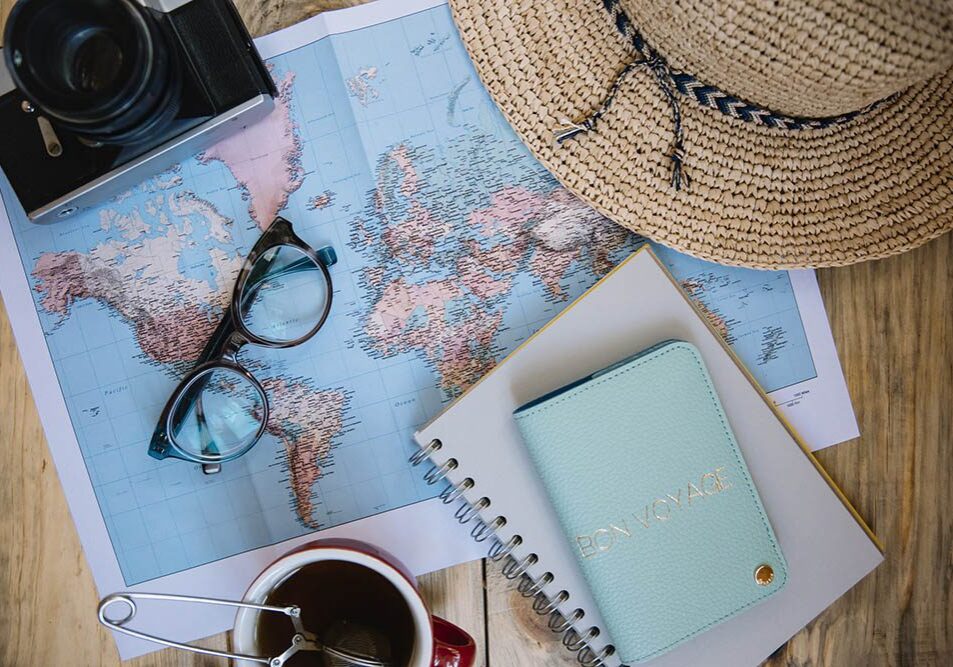 Travel preparations essentials. World map, cup of tea, vintage film camera, hat, passport, notebook and stylish glasses on the old rustic wooden table background, flat lay
